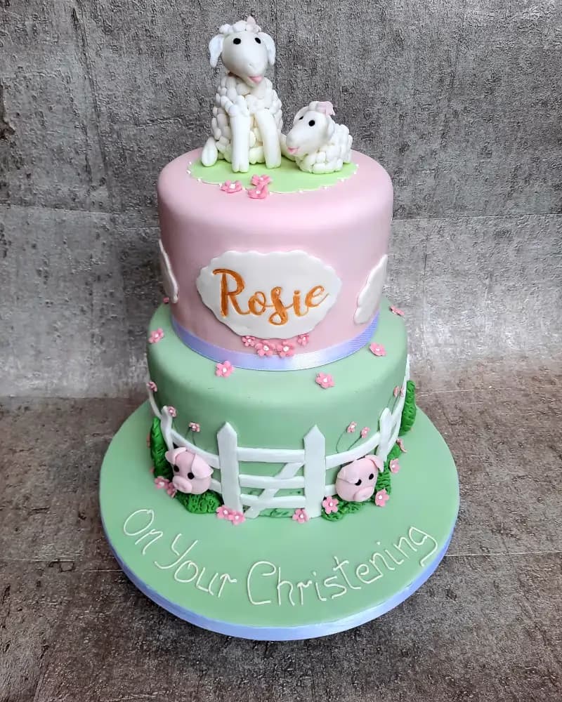A two-tier christening cake with a pink top tier and a light green bottom tier and base. The top tier features two white fondant sheep figures and a plaque with 'Rosie' written in gold. The bottom tier and base are decorated with a white picket fence, small pink pig figures, and tiny pink flowers. The cake base has 'On Your Christening' written in white script.