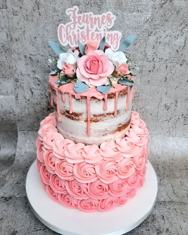 A two-tier christening cake with a 'Feurnes Christening' topper. The upper tier is a semi-naked cake with pink drip icing and sugar flowers, while the lower tier is covered in pink buttercream rosettes.