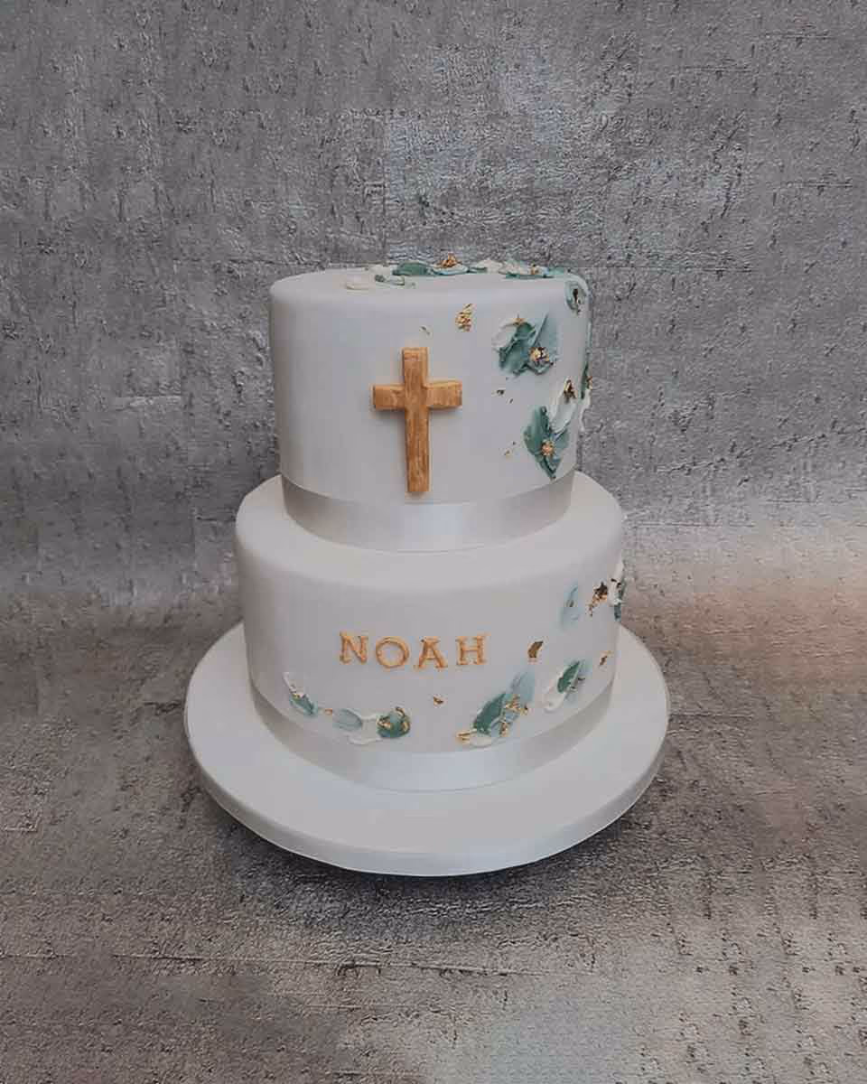A two-tier white christening cake featuring a gold cross on the top tier, with teal and gold abstract accents, and the name "NOAH" written in gold on the bottom tier.