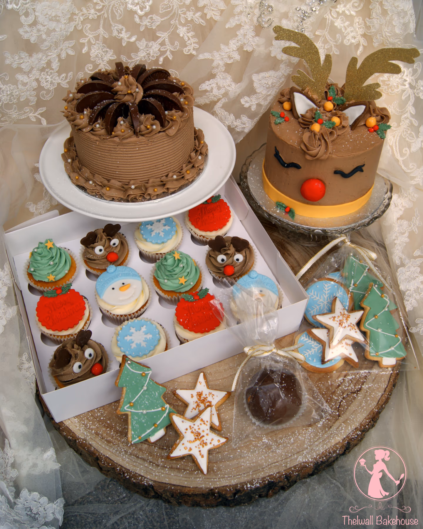 A festive display of Christmas baked goods on a wooden log slice, featuring a chocolate cake, a reindeer cake, a box of assorted Christmas-themed cupcakes, and decorated Christmas tree, star, and snowflake cookies along with a single chocolate truffle.