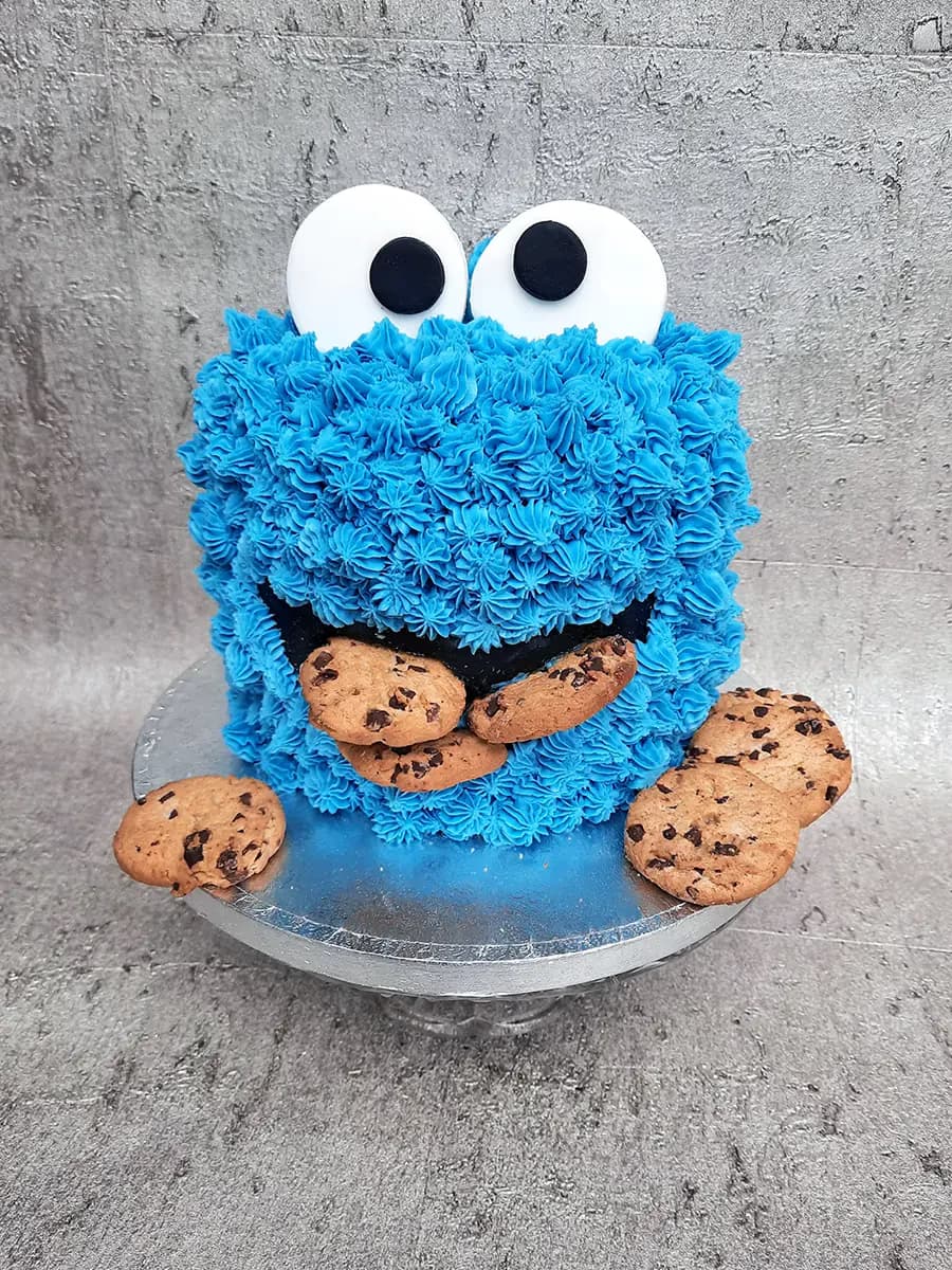 A vibrant blue Cookie Monster cake with large white and black eyes, decorated with several chocolate chip cookies around its mouth and on the silver cake stand.