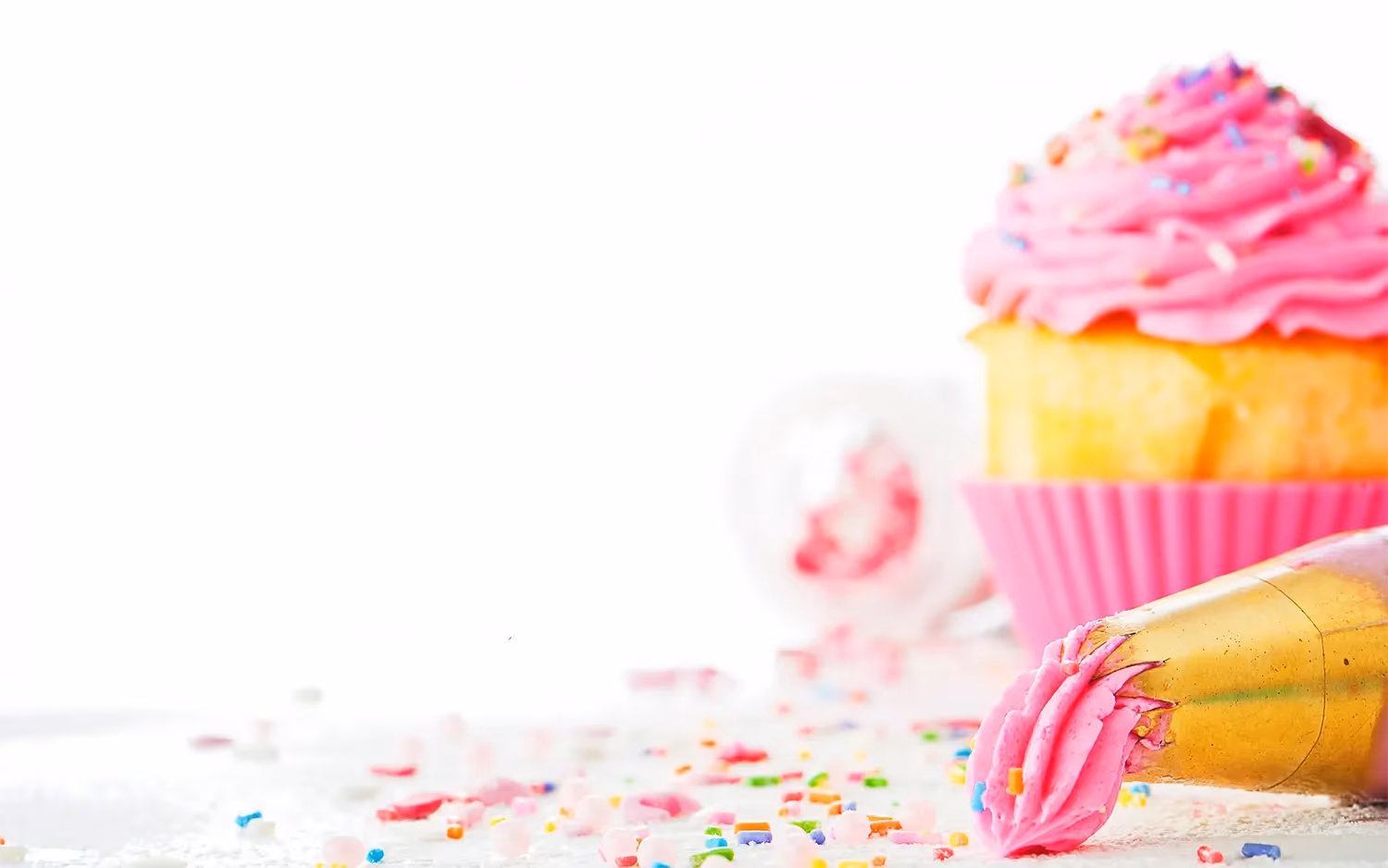A cupcake with bright pink frosting and colorful sprinkles in a pink liner, next to a gold piping bag with pink frosting, on a white surface scattered with more sprinkles.
