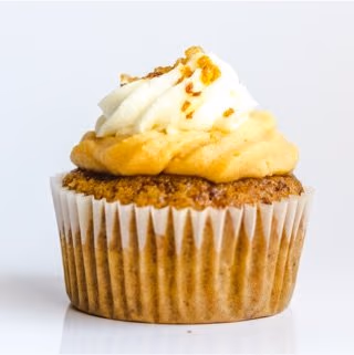 A single spice cake cupcake in a white paper liner, topped with a layer of light orange frosting and a swirl of white frosting, garnished with small orange pieces.