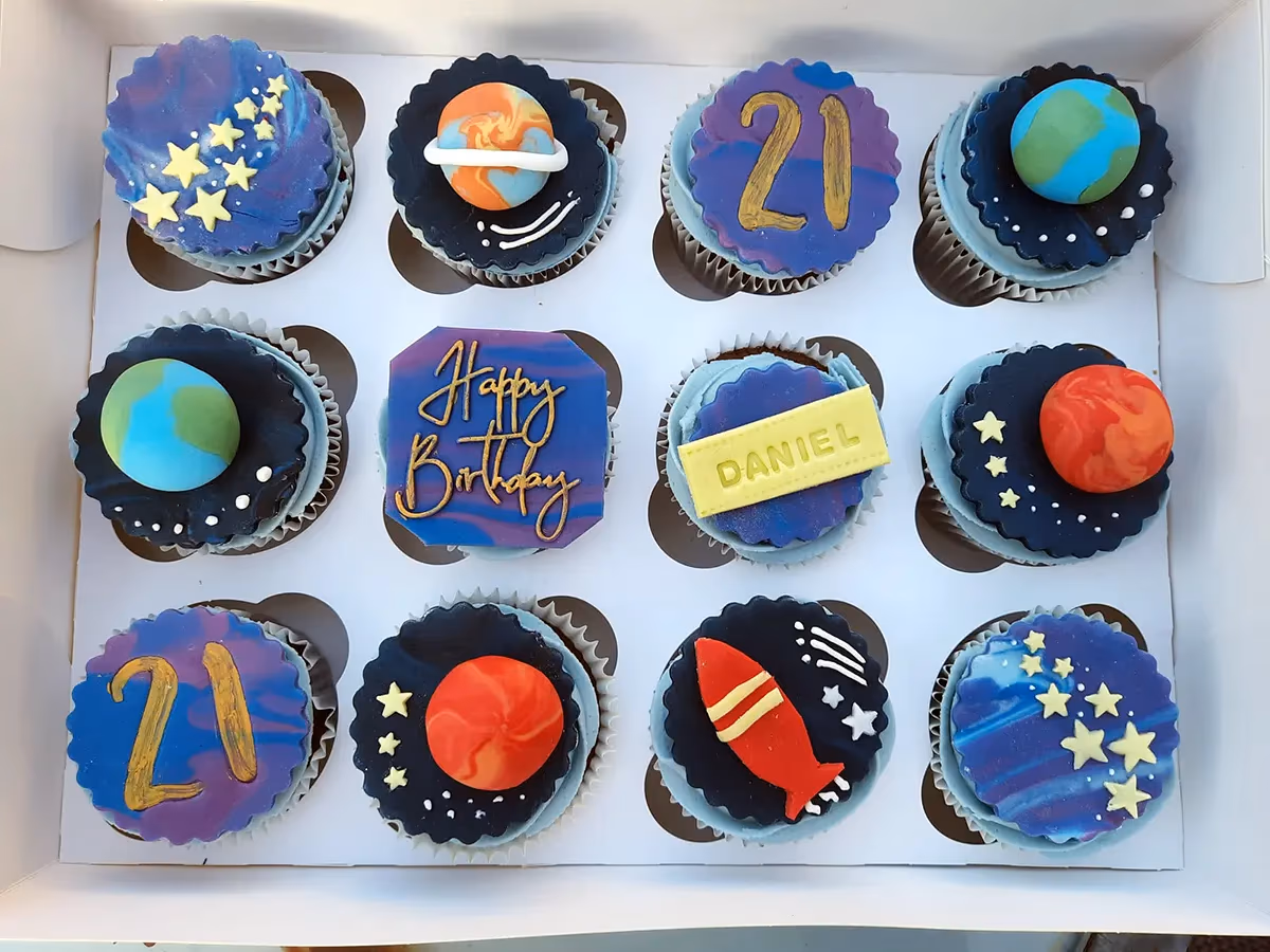 A box of twelve space-themed cupcakes for a 21st birthday, decorated with planets like Earth, Saturn, and Mars, stars, a rocket, 'Happy Birthday' in gold script, the number '21', and a yellow plaque with 'DANIEL'.
