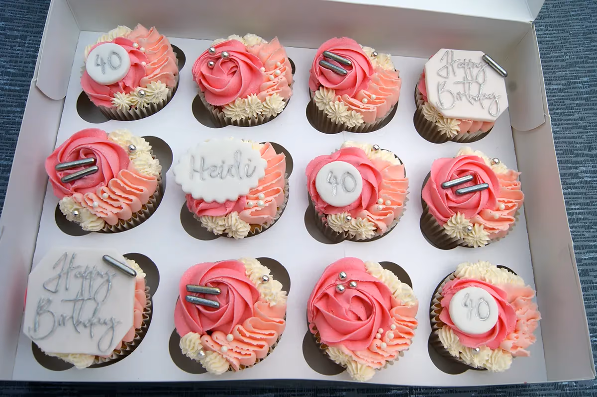 A white box containing twelve cupcakes, each decorated with swirls of pink and cream frosting. Some cupcakes feature white fondant toppers with '40', 'Heidi', or 'Happy Birthday' written in silver, while others have silver sprinkles or elongated silver sugar decorations.