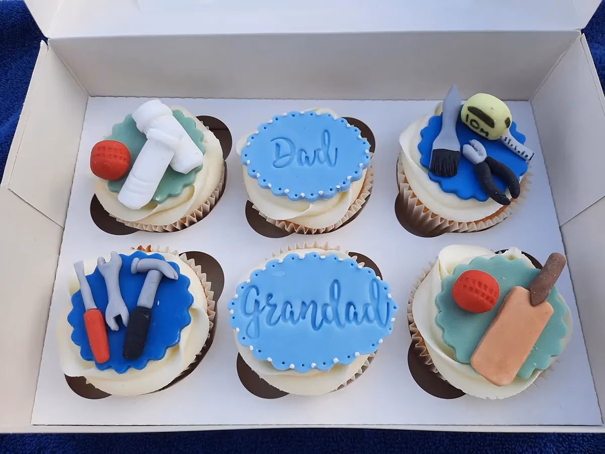 A white box containing six cupcakes with white frosting and various fondant toppers. Two cupcakes have blue oval toppers with 'Dad' and 'Grandad' written on them. Two cupcakes are cricket-themed, featuring bats, balls, and pads. The remaining two cupcakes are decorated with miniature tools, including a hammer, wrench, screwdriver, paintbrush, pliers, and a measuring tape.