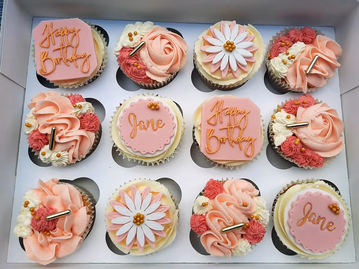 A white box containing twelve cupcakes, each decorated with various pink, white, and gold designs. Some cupcakes feature pink fondant toppers with 'Happy Birthday' or 'Jane' written in gold, while others have elaborate pink and white buttercream swirls, gold sprinkles, or delicate white and pink flower designs.