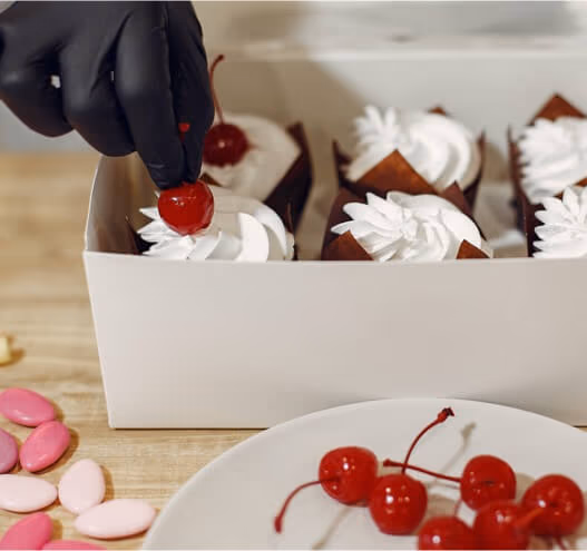 A gloved hand places a bright red cherry on a white-frosted cupcake, part of a box of freshly decorated cupcakes. More cherries and pink candies are visible on the wooden table.
