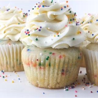 Close-up of a funfetti cupcake with a generous swirl of white frosting and colorful sprinkles, surrounded by other similar cupcakes.