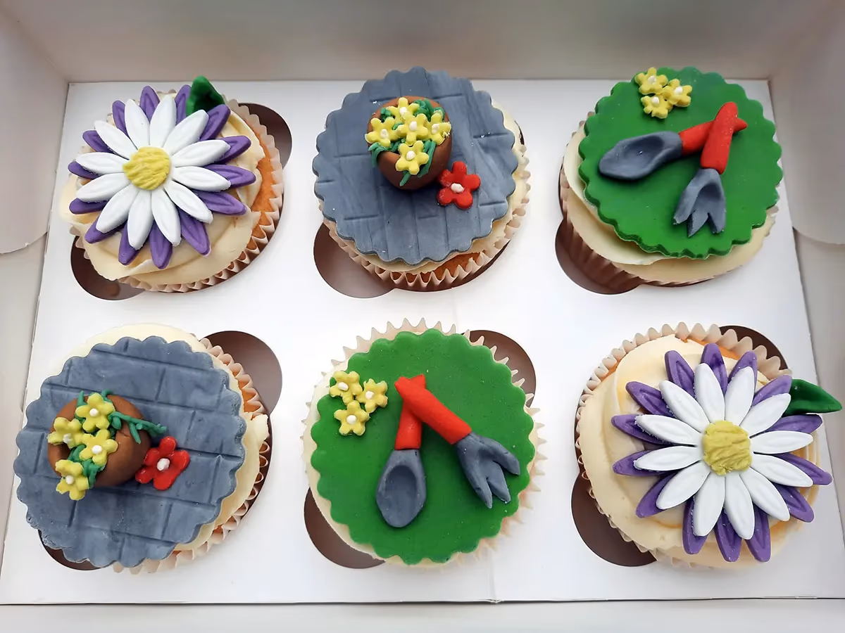 Six garden-themed cupcakes in a white box. Two cupcakes feature a large purple and white daisy, two have green frosting with miniature gardening tools and yellow flowers, and two have grey brick-patterned frosting with a potted plant and a red flower.