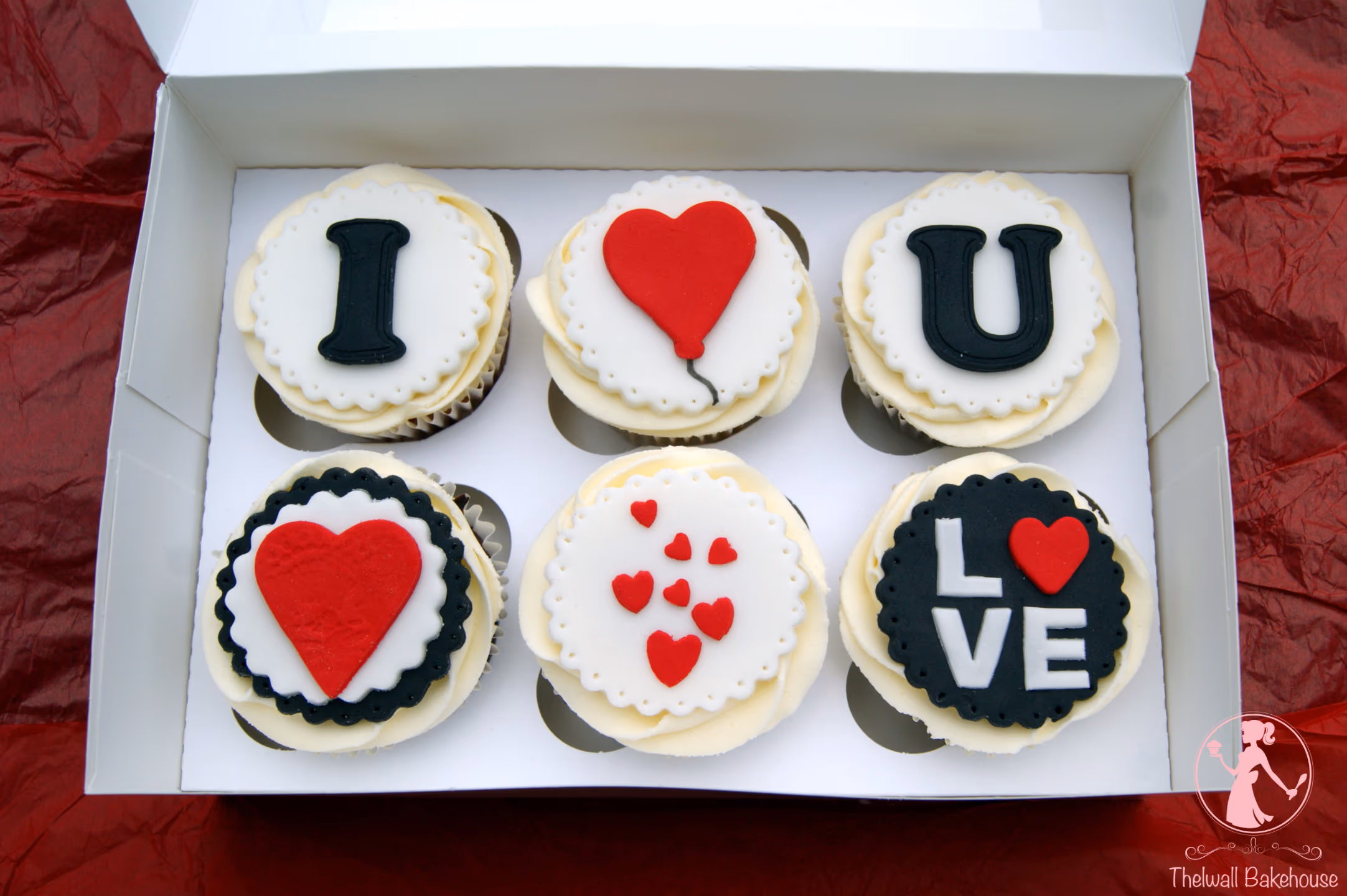 Six love-themed cupcakes in a white box, featuring white, black, and red fondant decorations that spell 'I LOVE U' and 'LOVE' with various heart designs.