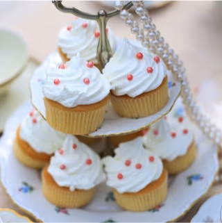 Miniature cupcakes with white frosting and small pink sprinkles arranged on a two-tier decorative stand, with a pearl necklace draped on the side.