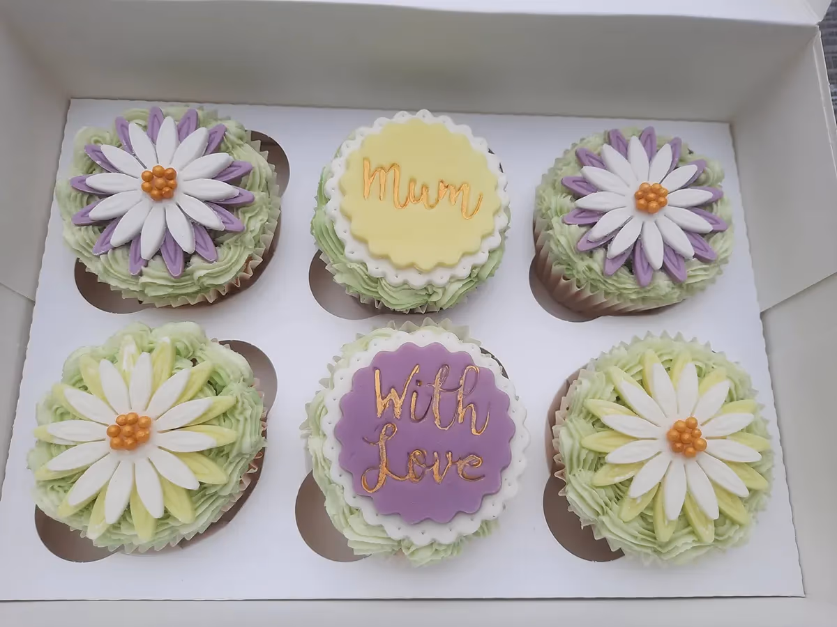 Six cupcakes in a white box, each with light green frosting. Four cupcakes are topped with daisy-like flowers with white petals and either purple or yellow accent petals. One cupcake has a yellow circular fondant topper with 'Mum' written in gold, and another has a purple circular fondant topper with 'With Love' written in gold.