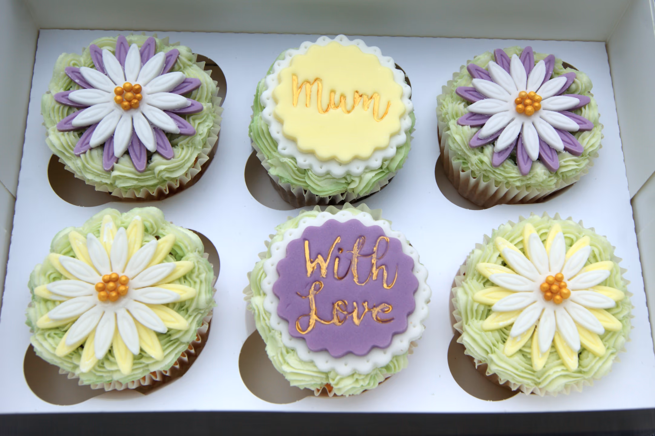 Six cupcakes in a white box, each with light green frosting. Two cupcakes are topped with purple and white fondant flowers, two with yellow and white fondant flowers, one with a yellow scalloped disc that says 'Mum' in gold script, and one with a purple scalloped disc that says 'With Love' in gold script.