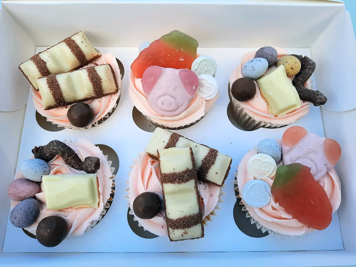 Six cupcakes with pink frosting in a white box, decorated with a variety of sweets and chocolates including Kinder Bueno bars, pink pig-shaped candies, gummy strawberries, mini chocolate eggs, and white chocolate bars.