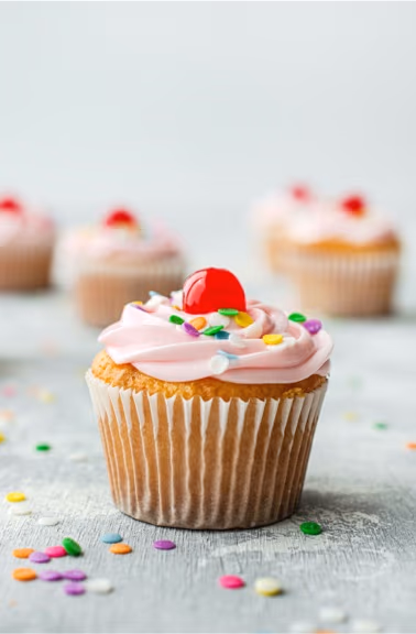A close-up of a cupcake with pink frosting, colorful sprinkles, and a red cherry, with more blurred cupcakes in the background on a light grey surface.