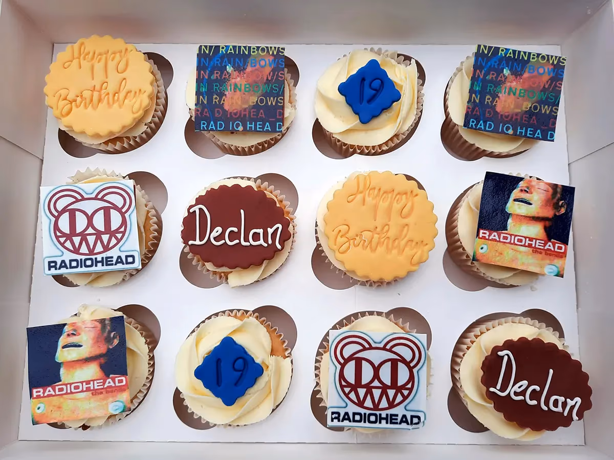 A dozen Radiohead-themed cupcakes in a white box, featuring various toppers including 'Happy Birthday', 'Declan', the number 19, the Radiohead bear logo, and album art from 'In Rainbows' and 'The Bends'.