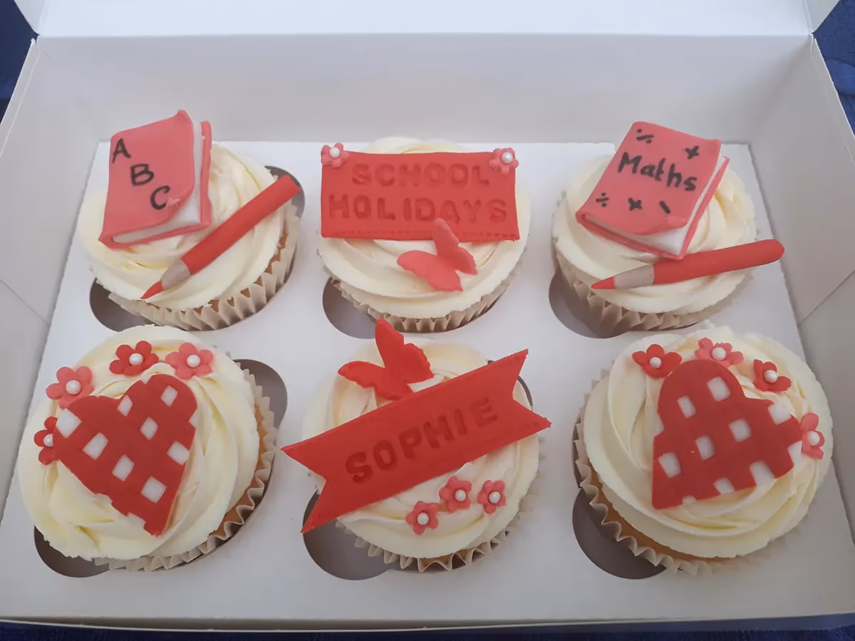 Six cupcakes with white frosting, decorated with various red school-themed toppers including books, pencils, 'SCHOOL HOLIDAYS' and 'SOPHIE' banners, butterflies, and checkered hearts, presented in a white box.