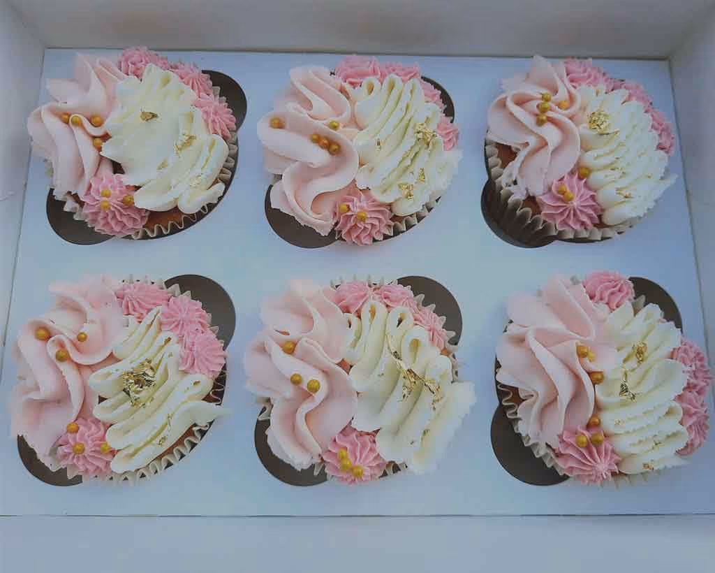 Six cupcakes arranged in a white box, each decorated with swirled pink and white frosting, small pink piped flowers, gold leaf flakes, and small gold spherical sprinkles.