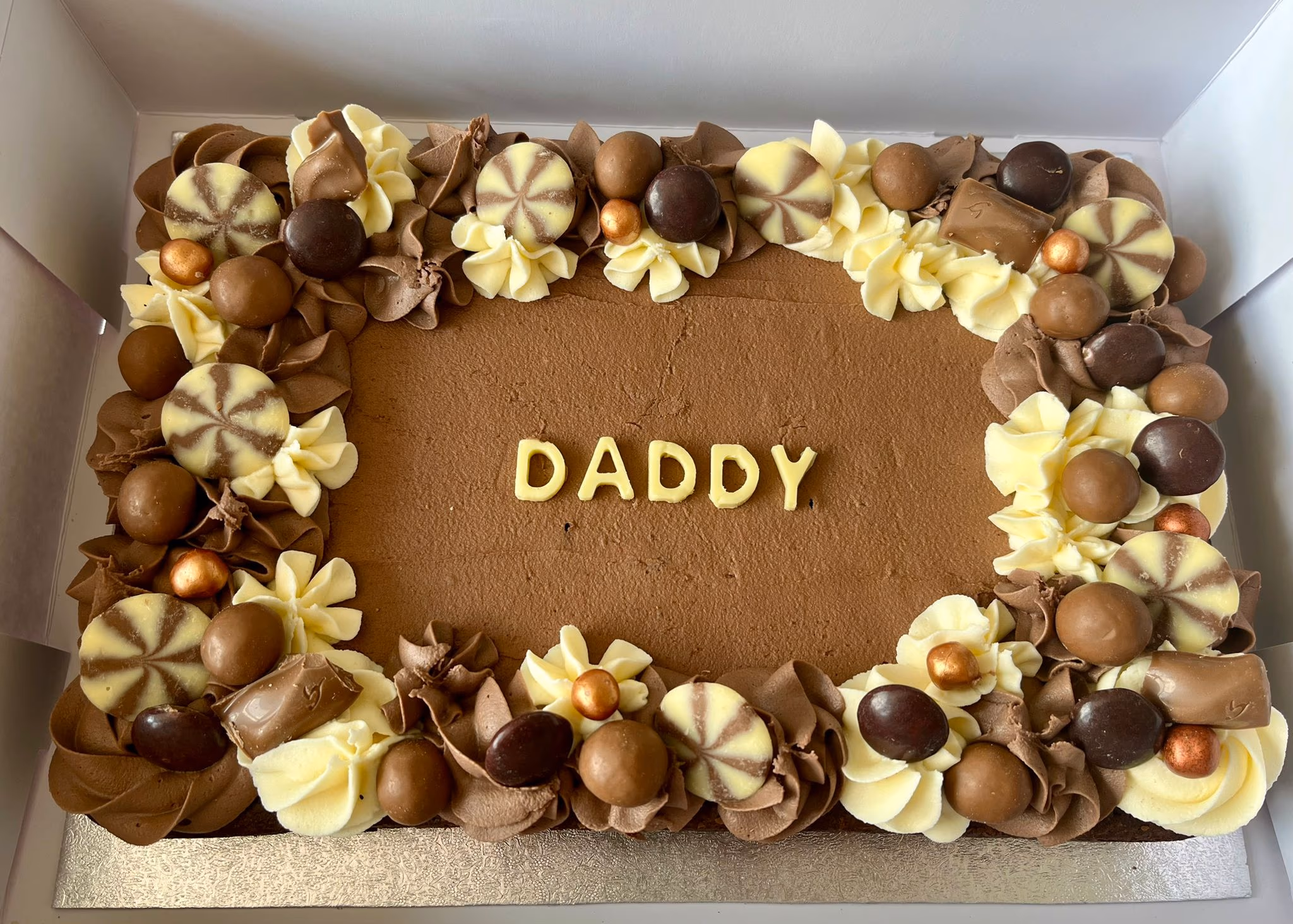 A rectangular chocolate cake with "DADDY" spelled out in yellow letters, surrounded by a decorative border of piped chocolate and cream frosting, various chocolate candies, and metallic bronze sprinkles.
