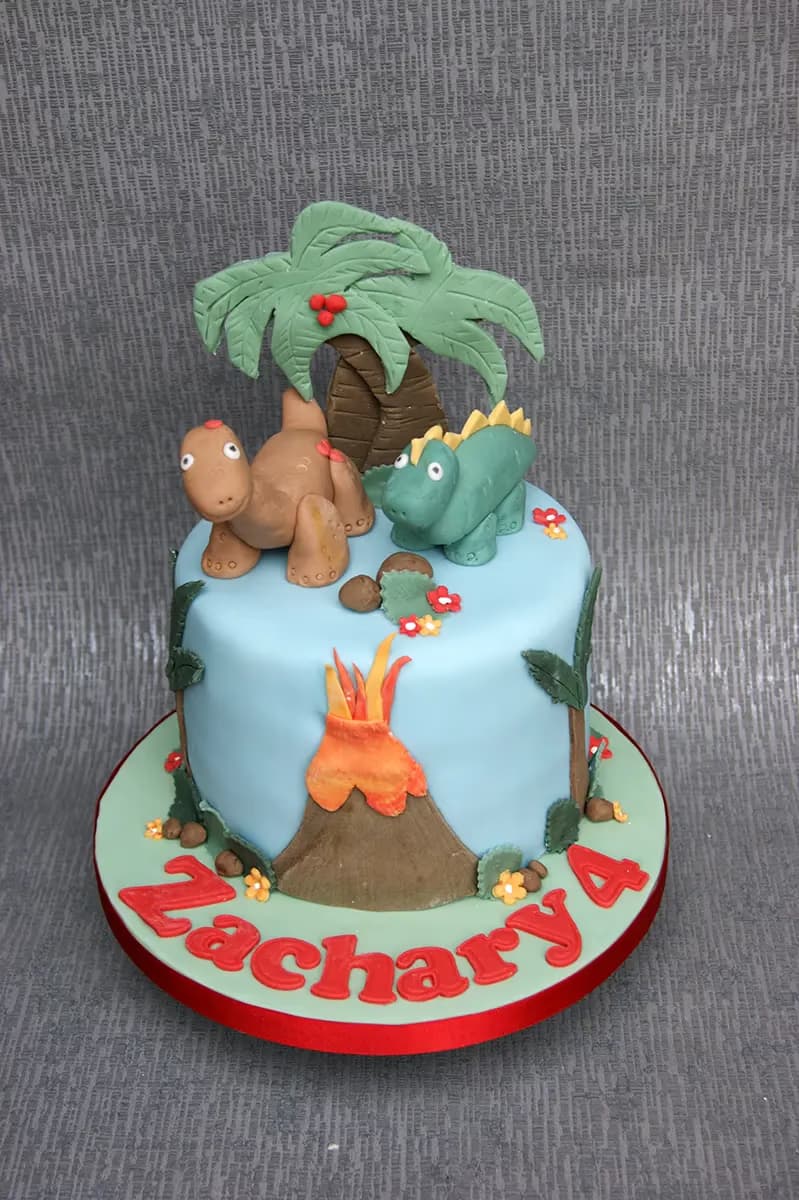 Single-tier light blue dinosaur-themed birthday cake for Zachary's 4th birthday, featuring two fondant dinosaurs, a palm tree, and a volcano with lava, all on a light green base with red lettering.