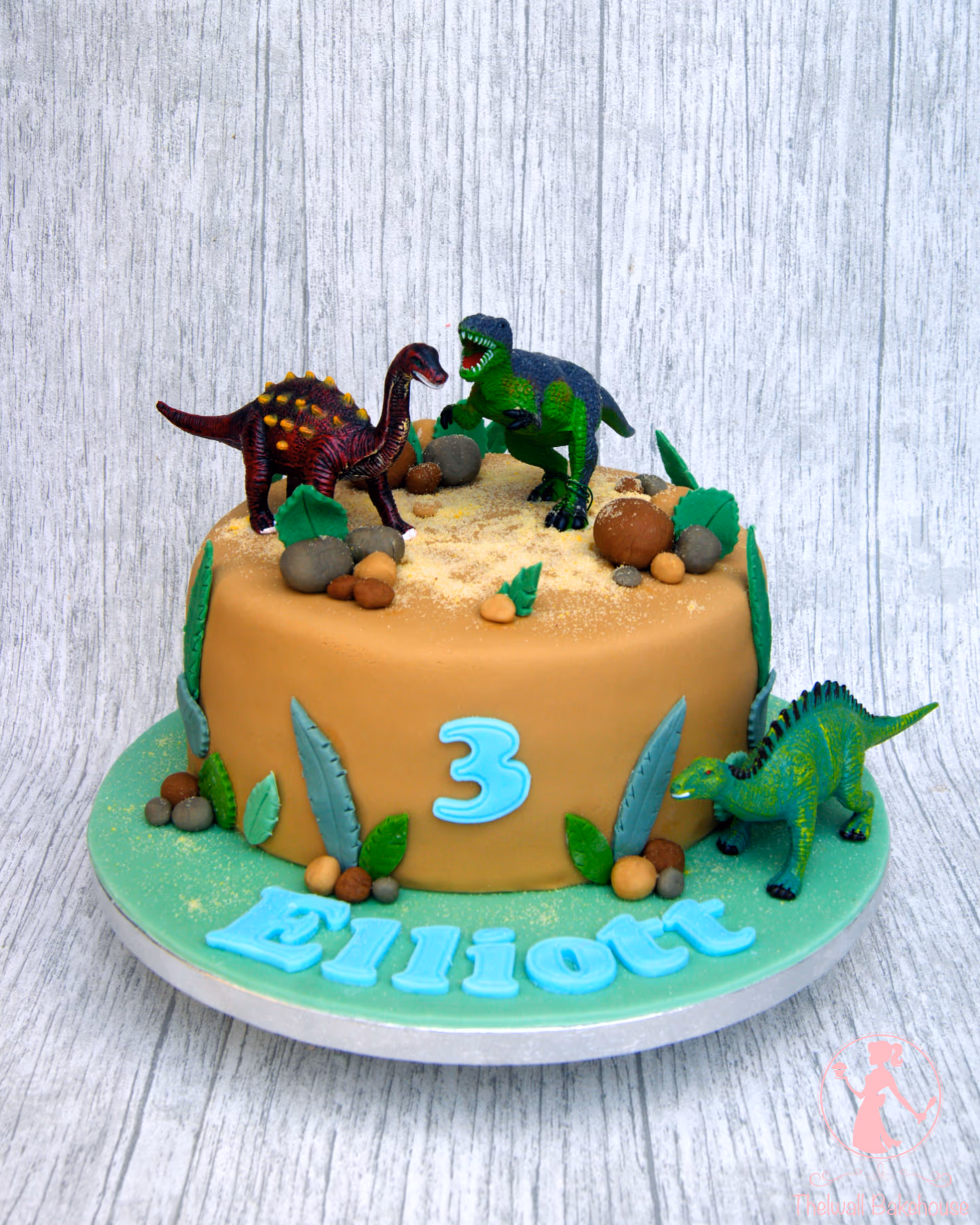 A round brown dinosaur-themed birthday cake for Elliot's third birthday, decorated with toy dinosaurs, edible rocks, sand, leaves, and a green cake board.