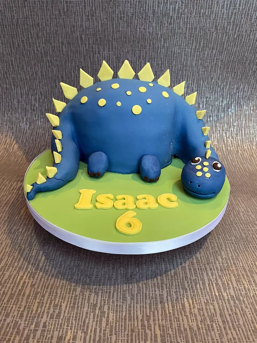 A blue dinosaur-shaped birthday cake with yellow spikes and polka dots, featuring the name 'Isaac' and the number '6' in yellow on a green base.