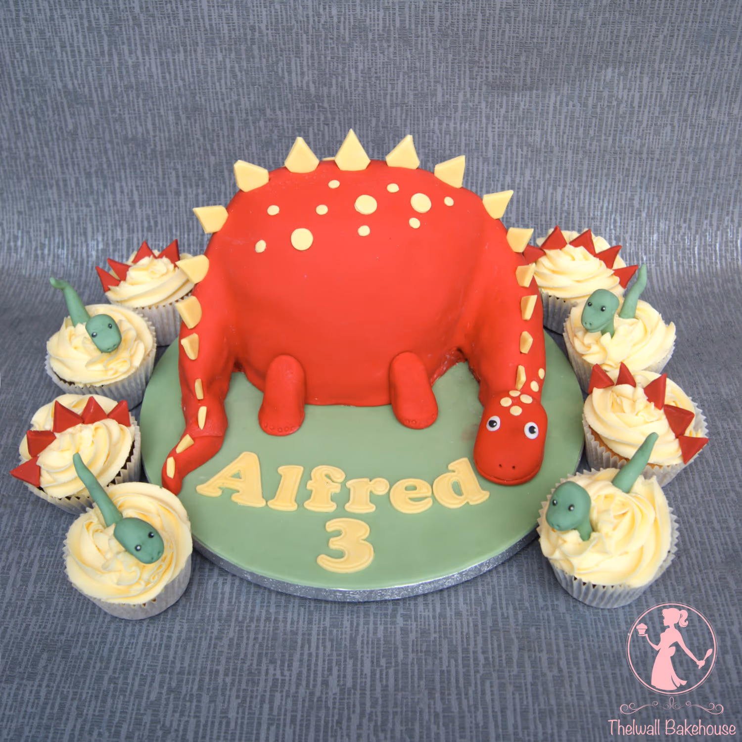 A vibrant red 3D dinosaur birthday cake with yellow spikes and spots, surrounded by matching cupcakes, some featuring green baby dinosaurs hatching and others with red triangular spikes. The green cake board is personalized with 'Alfred 3' in yellow fondant.
