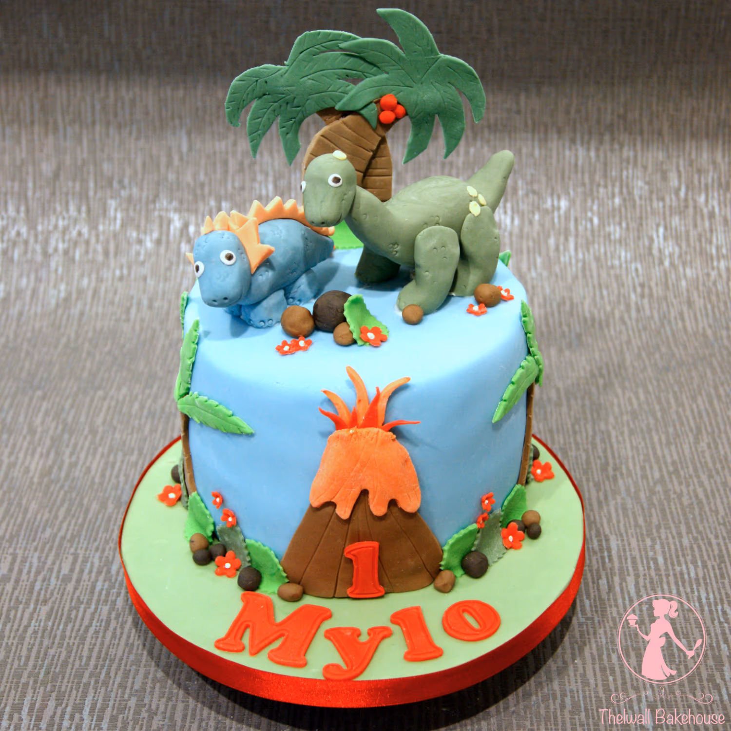 A light blue single-tier dinosaur-themed first birthday cake featuring a green dinosaur, a blue stegosaurus-like dinosaur, a palm tree, and an orange volcano with the number '1' on top, all on a light green base with the name 'Mylo' in red letters.