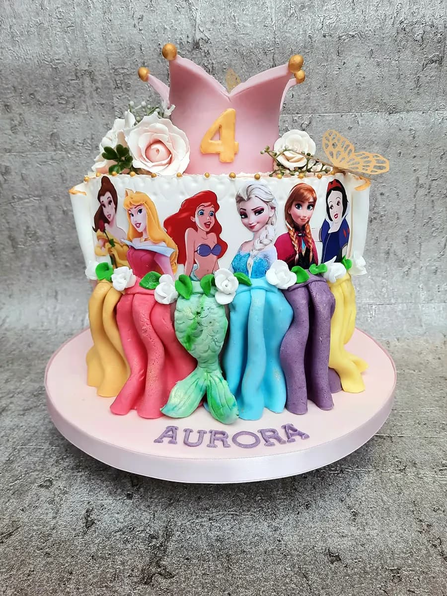 A vibrant Disney Princess birthday cake featuring images of Belle, Aurora, Ariel, Elsa, Anna, and Snow White, topped with a pink crown displaying the number 4, sugar flowers, a golden butterfly, and the name 'Aurora' written on the base.
