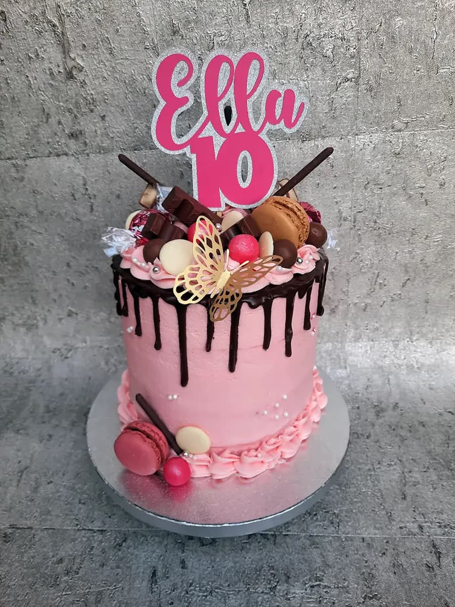 A tall pink drip cake decorated with a pink and silver 'Ella 10' cake topper, various chocolates, macarons, candies, a gold butterfly, and silver pearls, with dark chocolate ganache dripping down the sides and additional pink macarons and candies around the base.
