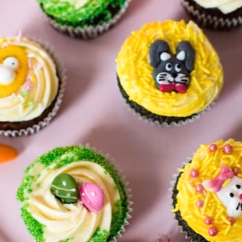 A top-down view of multiple Easter cupcakes on a pink background, featuring decorations like a black bunny on yellow frosting, a white bunny with a pink bow, a yellow chick, and colorful eggs and sprinkles.
