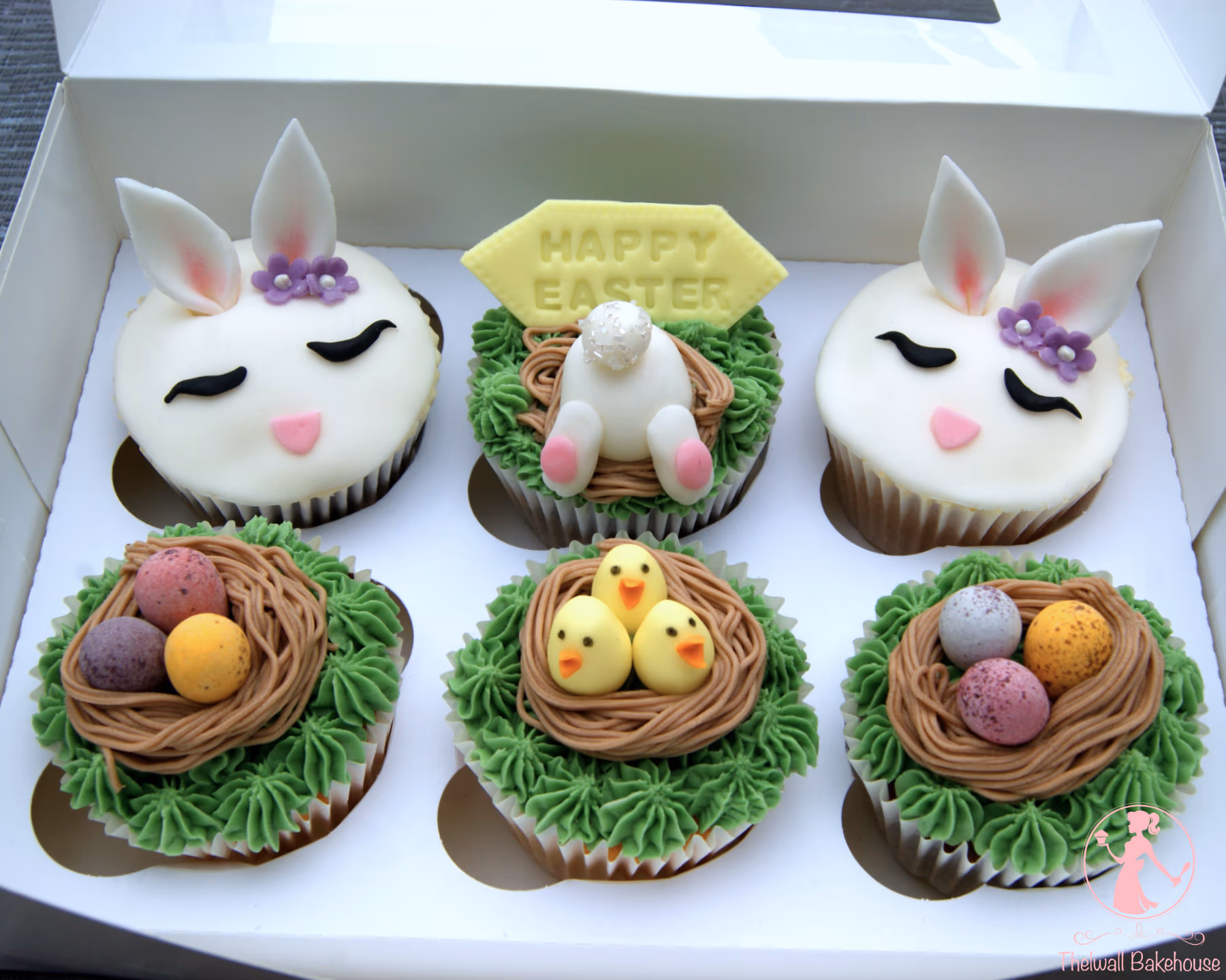 A box of six Easter-themed cupcakes. Two cupcakes are decorated as white bunny faces with pink noses, black closed eyes, and white ears, one featuring small purple flowers. One cupcake shows a white bunny's bottom with pink feet nestled in brown icing, surrounded by green frosting, with a yellow 'HAPPY EASTER' sign. The remaining three cupcakes have green grass-like frosting, each topped with a brown nest made of icing; one nest holds three yellow chick decorations, and the other two nests contain three speckled mini chocolate eggs in colors like pink, purple, white, and yellow.