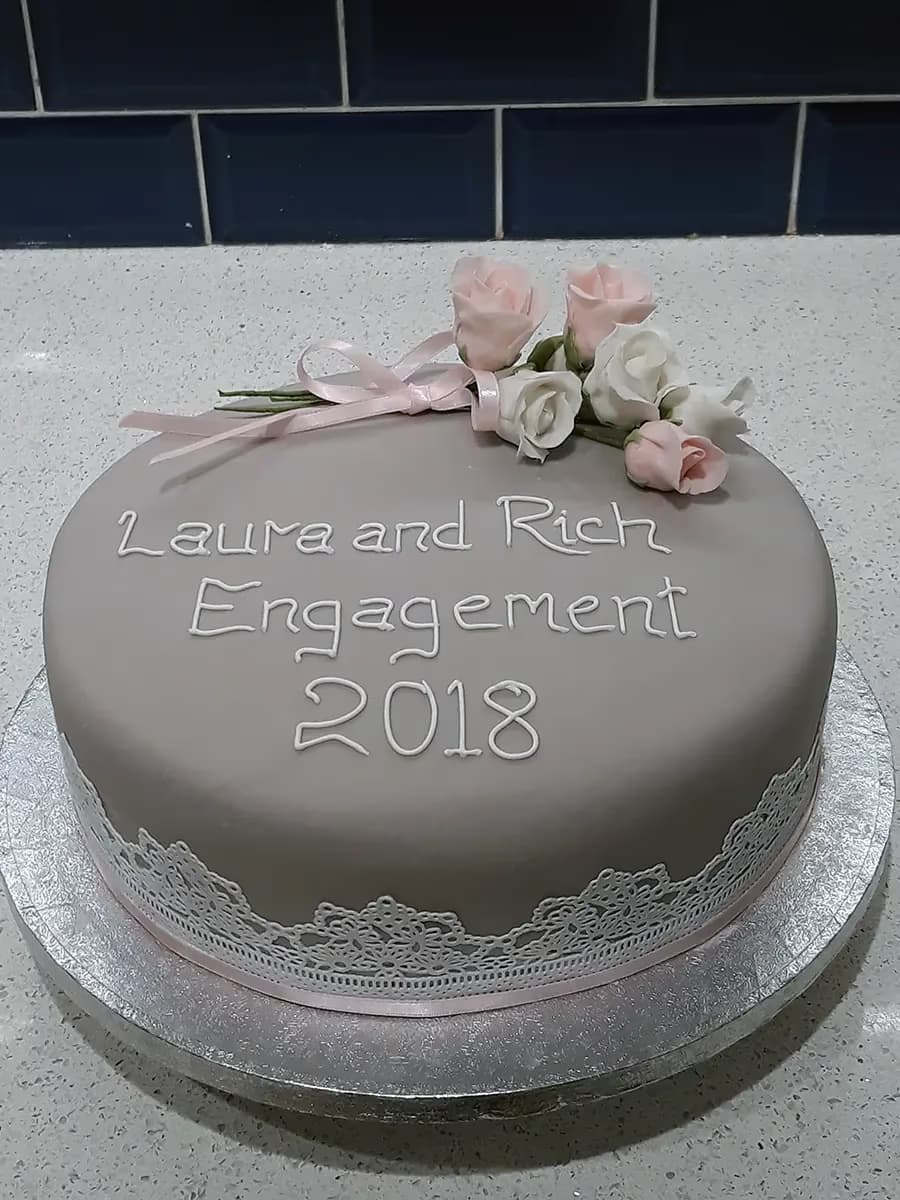 A round engagement cake with light grey fondant, decorated with white icing text 'Laura and Rich Engagement 2018', a cluster of pink and white sugar roses with a pink ribbon on top, and a white lace-like trim over a pink ribbon around the base.