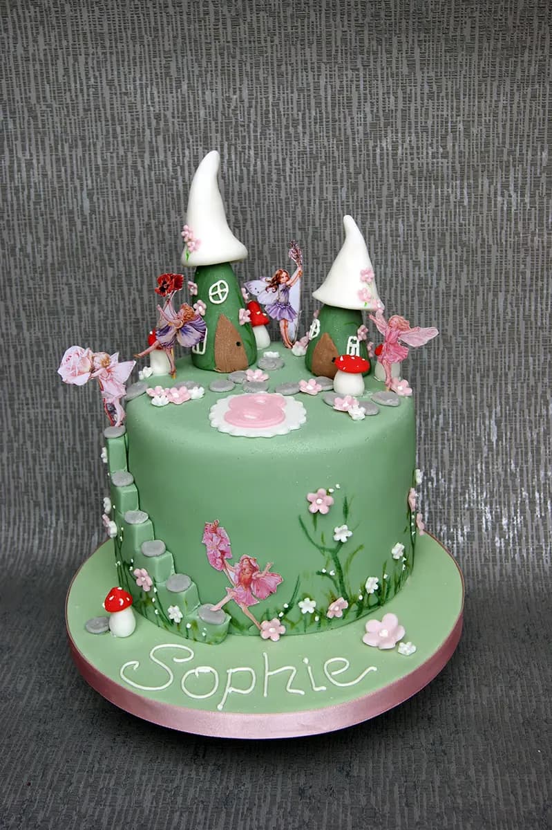 A round, single-tier birthday cake with light green fondant, decorated with a fairy garden theme. It features two mushroom-shaped houses with white roofs, several delicate fairy figures, small pink and white sugar flowers, grey steps, red and white toadstools, and the name 'Sophie' written in white icing on the front of the cake board. A pink number '8' is visible on top of the cake.