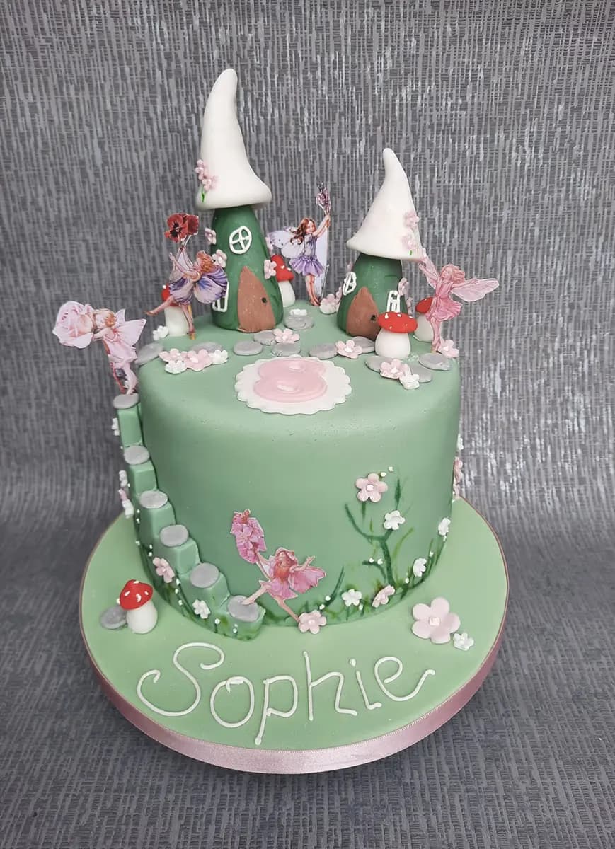 A light green, round fairy garden birthday cake featuring two fairy houses with white pointed roofs, various fairy cutouts, red and white mushrooms, white and pink flowers, and grey steps. The cake board has 'Sophie' written in white icing and there's a pink number '8' on top.