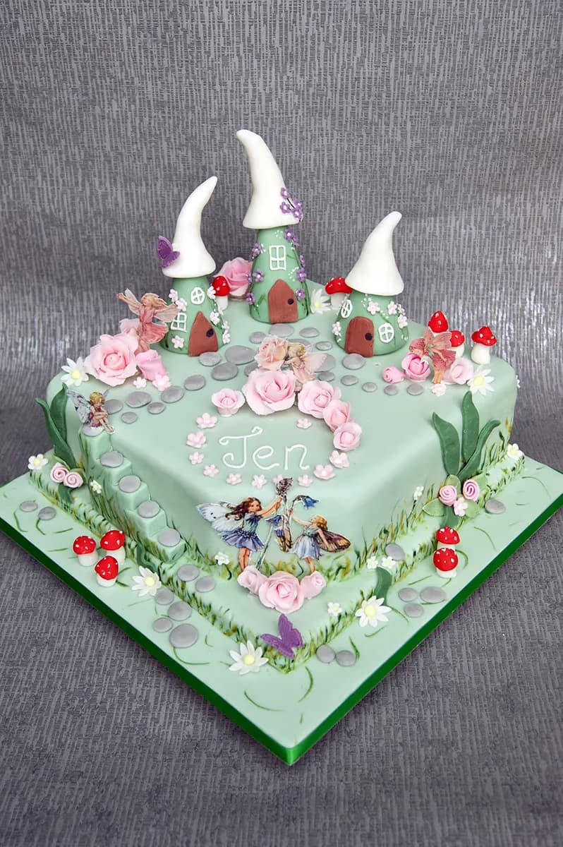 A square, light green fairy garden cake decorated with three gnome-like houses, pink roses, red and white mushrooms, fairy figures, and the name 'Jen' written in white icing.
