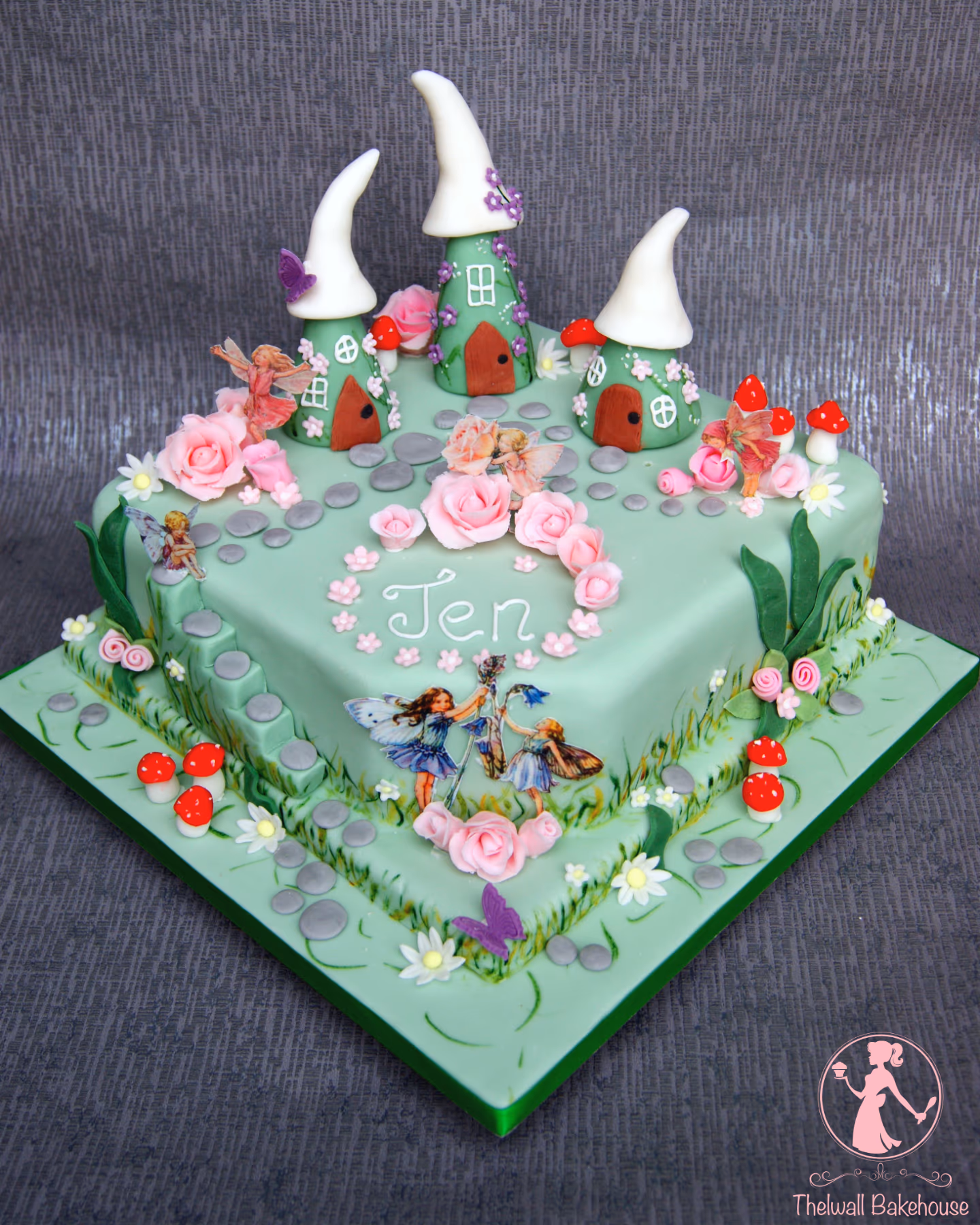 A whimsical two-tier square cake decorated with a fairy garden theme, featuring three green fairy houses with white roofs, sugar fairies, pink roses, mushrooms, and grey stones, with 'Jen' written on the top tier.