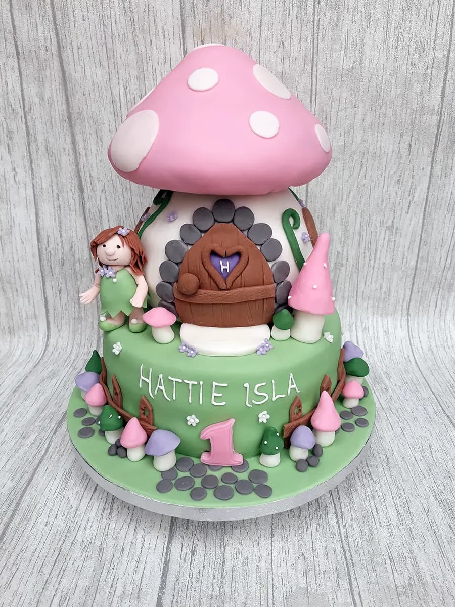 A two-tier first birthday cake designed as a fairy mushroom house. The top tier features a large pink mushroom cap with white polka dots over a white base with a brown door, stone details, and a small fairy figure. The bottom green tier is decorated with small fondant mushrooms and reads 'HATTIE ISLA' and a pink number '1'.