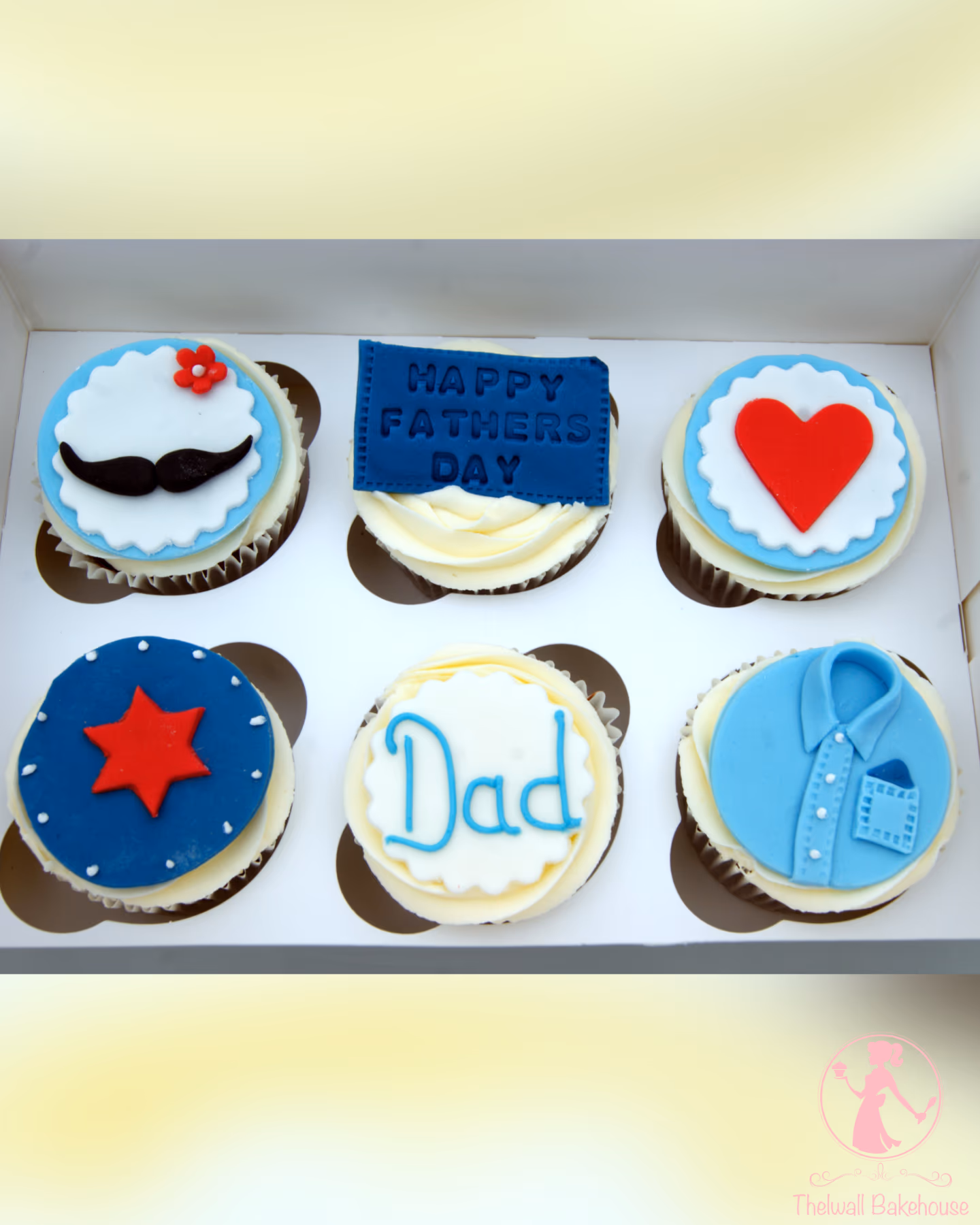 A white box holds six Father's Day cupcakes, each with a unique decoration: a black mustache with a small red flower, a blue banner reading 'Happy Fathers Day', a bright red heart, a red star on a blue circle, the word 'Dad' in blue script, and a light blue shirt with a pocket.