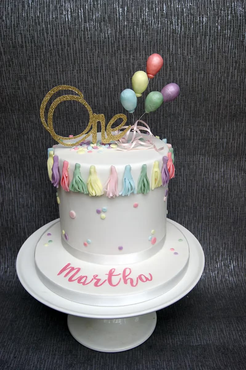 A single-tier white fondant first birthday cake decorated with a glitter gold 'One' topper, pastel balloons, pastel tassels, colorful sprinkles, and the name 'Martha' written in pink on the cake board.
