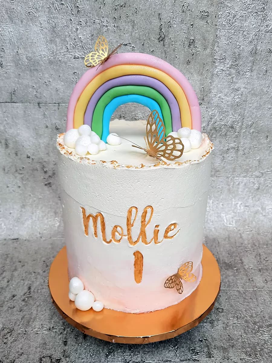 A single-tier white and pink ombre birthday cake for Mollie's first birthday, decorated with a pastel rainbow arch, white clouds, and gold butterfly accents.