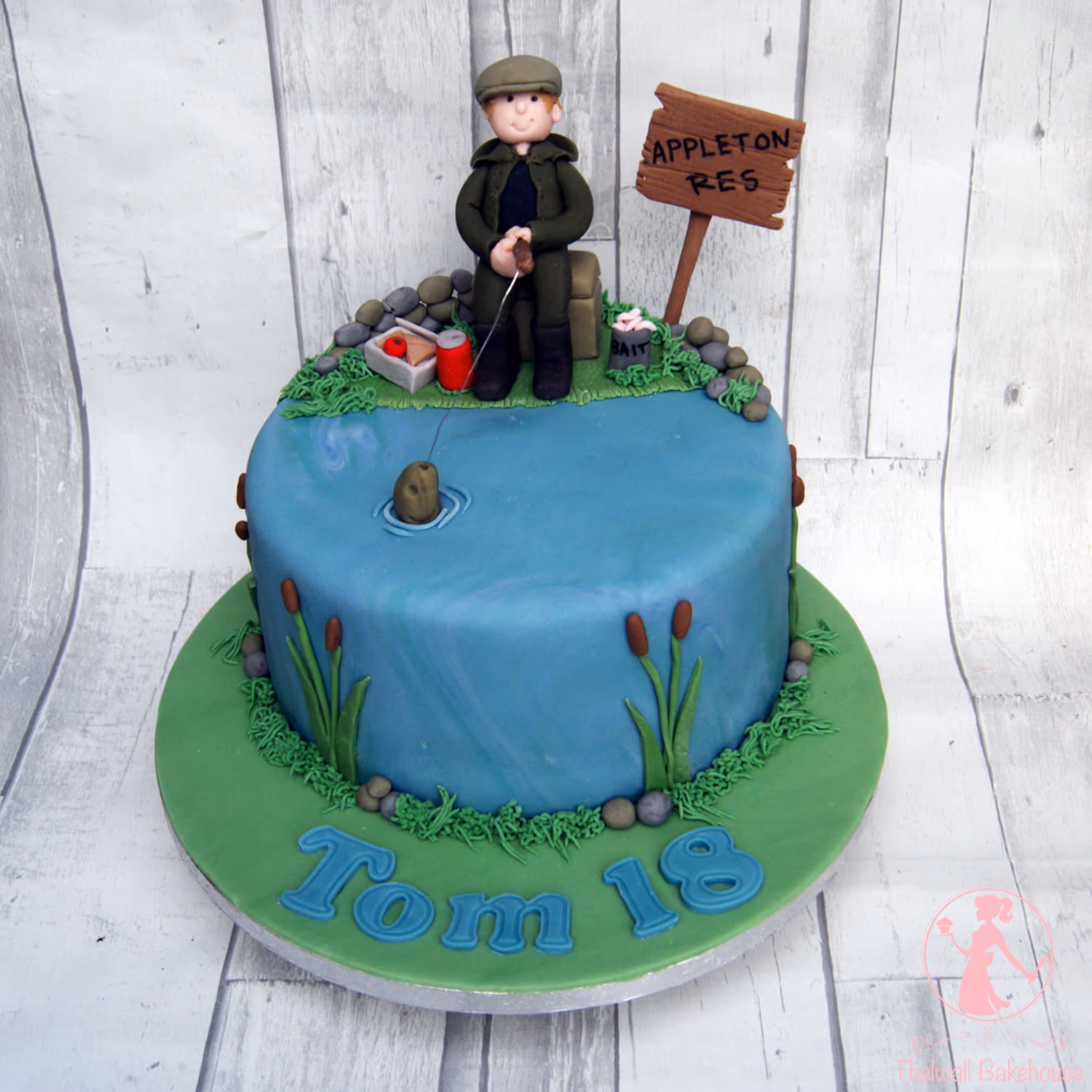A round, single-tier birthday cake decorated as a fishing scene, featuring a fondant figure of an angler in a green jacket and cap sitting by a blue pond with a fishing rod and bobber. The cake includes details like a 'APPLETON RES' sign, fishing bait, cattails, and 'Tom 18' written in blue fondant letters on the green cake board.