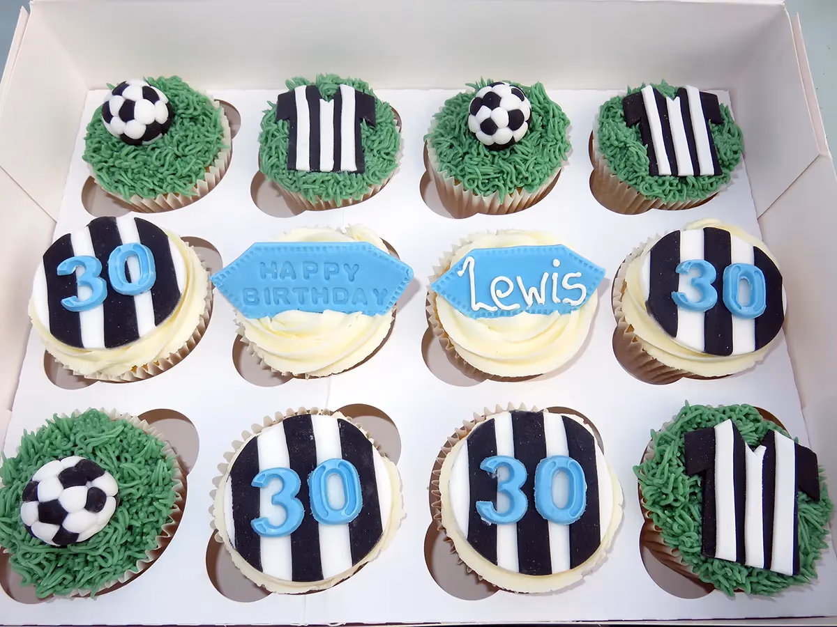 A box of 12 football-themed cupcakes for a 30th birthday, featuring green grass frosting with soccer balls and black and white striped jerseys, as well as cupcakes with black and white striped toppers and the number 30 in light blue, along with 'HAPPY BIRTHDAY' and 'Lewis' messages on blue plaques.