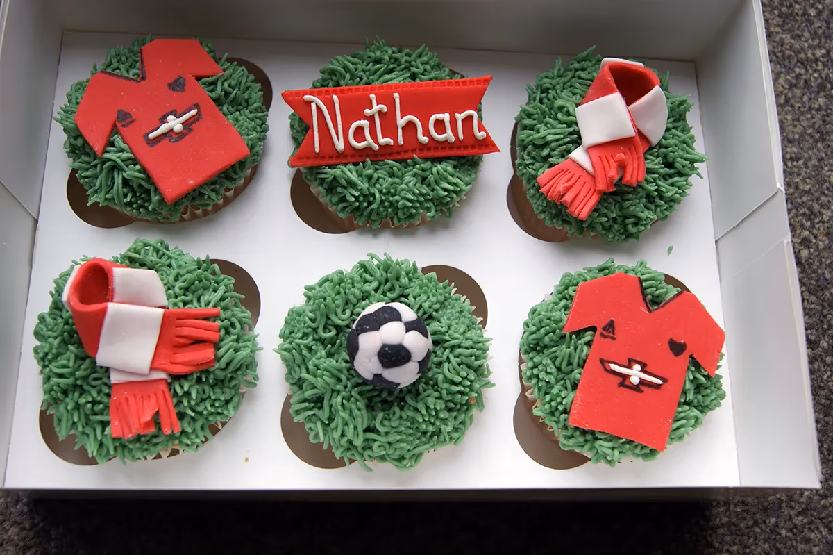 Six football-themed cupcakes in a white box, each topped with green grass-like frosting. Decorations include two red football jerseys, two red and white striped scarves, a black and white football, and a red banner personalized with the name 'Nathan'.