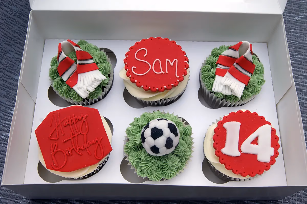 Six football-themed cupcakes in a white box, featuring green frosting with red and white scarves, a miniature football, and white frosting with red toppers displaying 'Sam', 'Happy Birthday', and the number '14'.