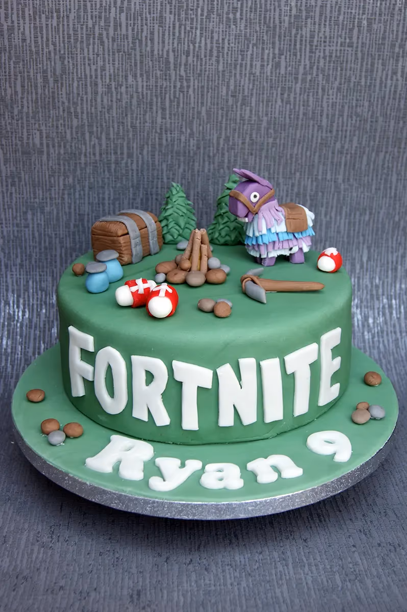 A green, round Fortnite birthday cake featuring a purple loot llama, treasure chest, campfire, pickaxe, mushrooms, and potions, with "FORTNITE" written on the side and "Ryan 9" on the base.