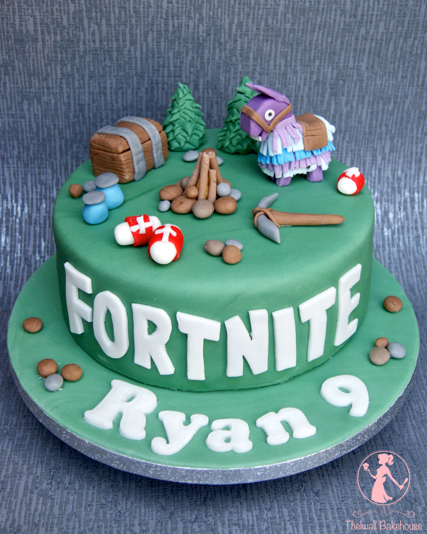 A two-tier green Fortnite-themed birthday cake featuring a purple loot llama, a treasure chest, green trees, a campfire with stones, a pickaxe, and red and white health items. The side of the top tier reads 'FORTNITE' and the bottom tier reads 'Ryan 9' in white letters.