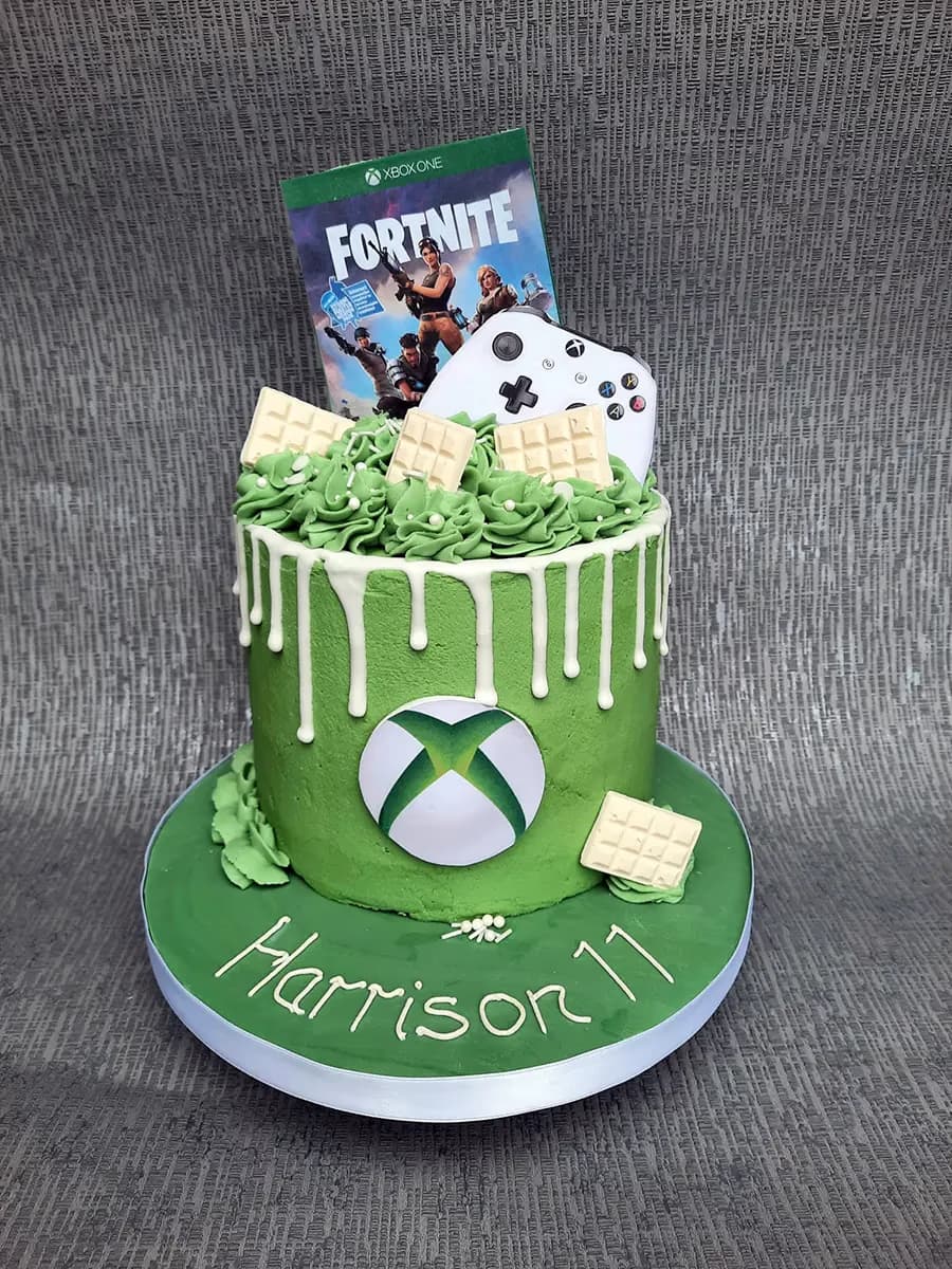 A vibrant green birthday cake with white drip icing, featuring an Xbox logo on the side and topped with a Fortnite game box, an Xbox controller, and white chocolate squares. The green cake board is decorated with white icing spelling 'Harrison 11'.
