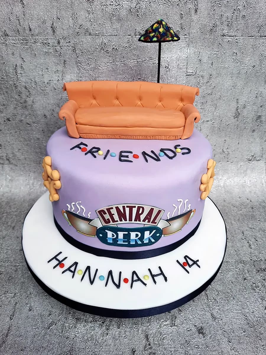A two-tier Friends-themed birthday cake with a light purple base, topped with an orange couch and a miniature stained-glass lamp. The cake features the "FRIENDS" logo with colored dots, the "CENTRAL PERK" logo, and is personalized with "HANNAH 14" on the white cake board.